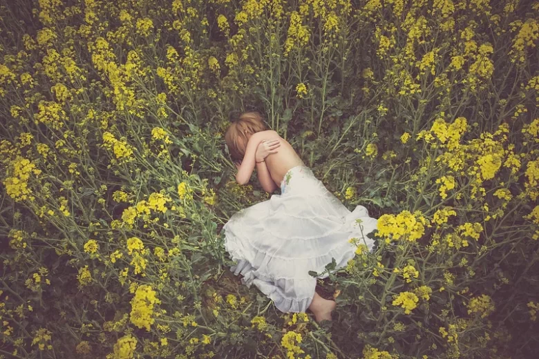 Jovem mulher, loira, deitada num campo de flores amarelas, em posição fetal, a dar um abraço ao seu corpo